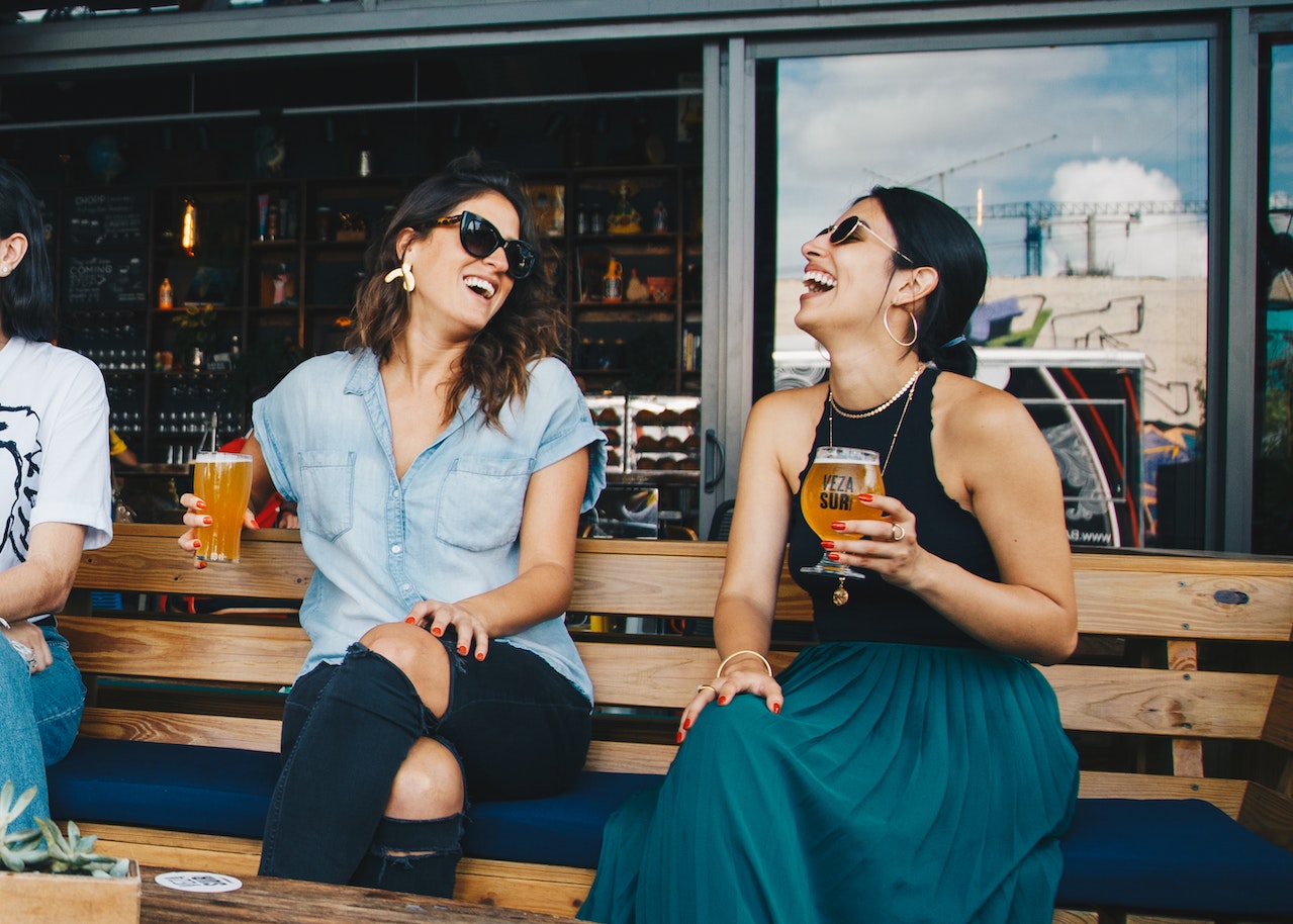 Two women laughing while enjoying a beer on a wooden bench.