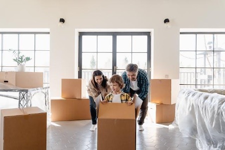 A family of three smiles while opening a box in a bright, spacious room with boxes around